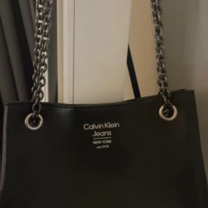 Svart axelväska från Calvin Klein - Snygg svart axelväska från Calvin Klein Jeans med kedjehandtag. Perfekt för en stilren look och rymmer det mesta du behöver ha med dig. Väskan har en enkel och elegant design med märkets logga framtill.
