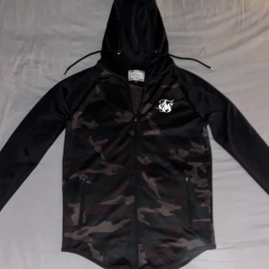 SikSilk Zipper limiterad - En SikSilk muscle zipper med färgerna camo och svart med vitt tryck. Storleken är M och sitter hyfsat tajt med töjbart material (perfekt för träning men även vid vanliga aktiviteter). Den är endast använd vid ett enstaka tillfälle och är i nyskick. Nypris: cirka 1000kr