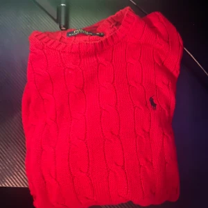 Röd stickad tröja från Ralph Lauren - Cable-Knit Cotton Jumper.    Säljer en snygg röd stickad tröja från Ralph Lauren med kabelstickat mönster och den klassiska loggan på bröstet. Perfekt för kyliga dagar och en stilren look.