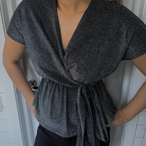Glittrig omlottopp med knytband - Snygg glittrig omlottopp i svart/silvrigt glitter, korta ärmar och knytband i midjan🖤Perfekt för en fest eller tillställning😊 köpt secondhand i london för 200, ej använd av mig- så därför priset 
