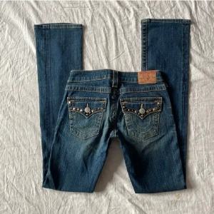 Lågmidjade jeans från True Religion - Snygga blå jeans från True Religion med dekorativa detaljer på fickorna. Jättebra skick! Midjemått rakt över 34-38 (stretch), grenhöjd 18, benöppning 17.5, innerbenslängd 85, lår 20. 