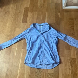 Blå skjorta med palmmönster från Denim Day - Snygg blå skjorta från Denim Day med små palmmönster. Skjortan har långa ärmar och knappar framtill. Perfekt för en avslappnad stil med en touch av sommarvibbar. 🌴