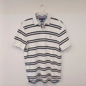 Randig pikétröja från Tommy Hilfiger - Snygg randig piké från Tommy Hilfiger med marinblå ränder. Tröjan har en klassisk passform och korta ärmar, perfekt för en stilren look. Den ikoniska loggan är broderad på bröstet. Perfekt för både vardag och avslappnade tillfällen. Den är helt oanvänd och priset är inte hugget i sten😀