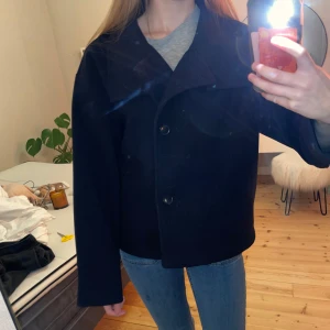 Marinblå kappa med knappar - Snygg svart kappa med stora knappar framtill. Jackan har en klassisk design med bred krage och långa ärmar. Perfekt för höst och vår.