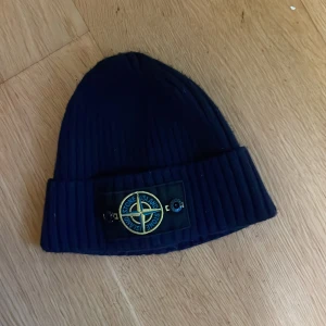 Mörkblå ribbad mössa från Stone Island - Snygg mörkblå mössa från Stone Island med ribbad design och det ikoniska märket framtill. Perfekt för att hålla värmen under kyliga dagar.   