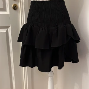 Svart volangkjol - Snygg svart kortkjol med volanger. Kjolen har en elastisk midja för bekväm passform och är perfekt för att ge en trendig look.