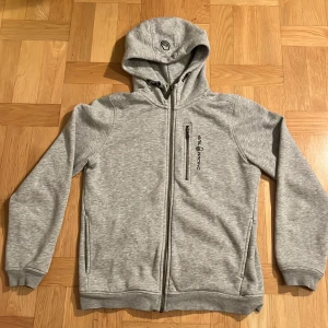 Grå Sail Racing hoodie med dragkedja  - Säljer en grå hoodie med dragkedja och huva. Den har en broderad logga på bröstet och en praktisk ficka med dragkedja. 