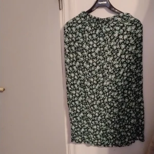Blommig kjol från Lindex - Säljer en fin blommig kjol från Lindex i storlek 170. Kjolen har en svart bas med ett vackert blommönster i grönt och vitt. Den har en slits framtill som ger en extra detalj till designen.