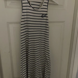 Randig ärmlös klänning från Marine - Säljer en stilren vit och marinblå randig klänning från Marine. Den är ärmlös och har en broderad detalj på bröstet. Perfekt för en avslappnad dag.