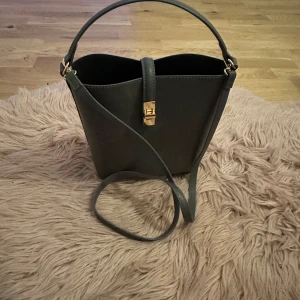  H&M  handväska med axelrem - Elegant handväska i skinn med justerbar axelrem och ett stilrent spänne i guld. Perfekt för en chic och minimalistisk look.