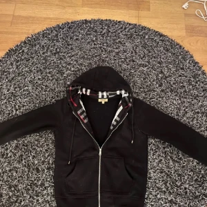 Svart huvtröja från Burberry - Säljer en stilren svart huvtröja från Burberry med klassiskt rutigt mönster på insidan av huvan. Tröjan har dragkedja framtill och är tillverkad i bomull. Säljer för den är för liten för mig men nydligen inköpt och knappt använd 