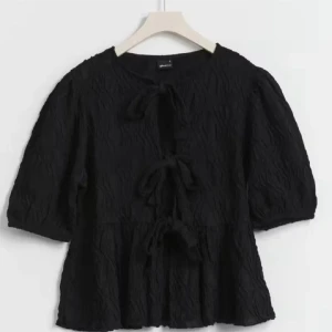 Svart blus med knytning - Elegant svart blus med knytning framtill och trekvartsärmar. Blusen har en texturerad yta som ger en unik look. Perfekt för en stilren och chic outfit.