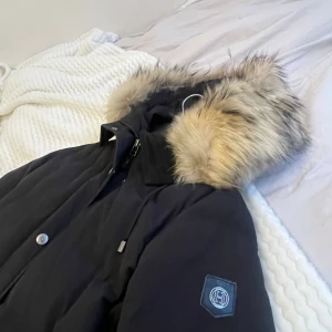 Svart dunjacka från Hollies - Säljer en svart dunjacka från Hollies med en stor, fluffig pälsfodrad huva. Jackan har dragkedja och knappar framtill samt en snygg logga på ärmen. Perfekt för kalla vinterdagar.