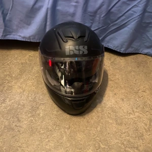 Svart hjälm från BSS - Säljer en svart hjälm från BSS med visir. Hjälmen har en stilren design och är perfekt för motorcykelentusiaster. Den är utrustad med ventilationsöppningar för extra komfort.