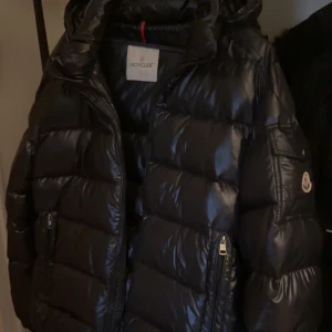 Svart pufferjacka från Moncler - Snygg svart pufferjacka från Moncler med glansigt material och dragkedja. Perfekt för kyliga dagar med sin vadderade design och huva. Storlek M-L 