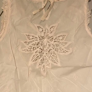 Vit ärmlös blus med spetsdetaljer - Säljer en söt vit ärmlös blus från Odd Molly med vackra spetsdetaljer och knytning i nacken. Blusen har en broderad design på ryggen och volangkant vid ärmhålen. Perfekt skick