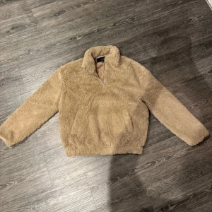 Beige teddytröja från Vero Moda - Mysig beige teddytröja från Vero Moda med dragkedja framtill. Perfekt för kyliga dagar med sin fluffiga och varma design. Tröjan har långa ärmar och en hög krage för extra värme.