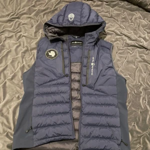 Mörkblå sail racing väst - Snygg mörkblå dunväst från sail racing med huva och dragkedja. Västen har ett stilrent märke på bröstet och är perfekt för kyligare dagar,den är i nyskick och har väldigt skön material