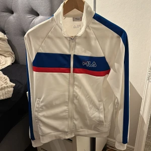Vit kofta från Fila - Säljer en vit träningsjacka från Fila med blå och röda detaljer. Koftan har dragkedja framtill och långa ärmar. Perfekt för en sportig look! 