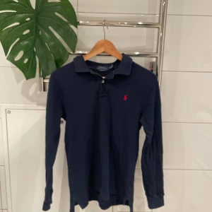 Mörkblå pikétröja från Ralph Lauren - Säljer en klassisk mörkblå pikétröja från Ralph Lauren med lång ärm och den ikoniska loggan broderad i rött på bröstet. Perfekt för en stilren och avslappnad look.
