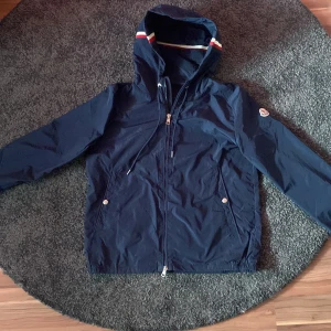 Moncler, är öppen för bud !  - Snygg mörkblå vindjacka från Moncler med huva och dragkedja. Den har en stilren design med ränder i huvan och Moncler-logga på ärmen. Säljer min Moncler jacka i o med att den har blivit för liten för mig. Köpte den ny för 7000 kr, har haft den i 1 å. Den är i 8/10 skick. ”Givetvis äkta” Storlek s