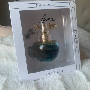Luna parfym från Nina Ricci - Elegant parfymflaska formad som ett äpple med en skimrande blå och guld färg. Flaskan har en gyllene spraypump dekorerad med blad. Förpackningen är stilren med en vit ram och svart text.