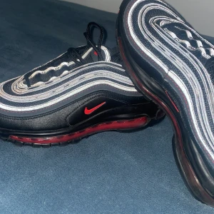 Nike Air Max 97 i svart och röd - Snygga Nike Air Max 97 sneakers i svart med röda detaljer. Skorna har en ikonisk vågig design med synlig Air-enhet i sulan för extra komfort. Perfekta för en sportig look. Skorna kan ser ut och vara på en bra form.