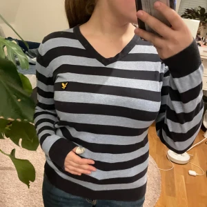 Randig tröja från Lyle & Scott - Säljer en snygg randig tröja från Lyle & Scott i svart och grått. Tröjan har en v-ringad hals och ett broderat gult emblem på bröstet. Perfekt för en stilren look!