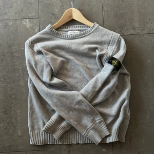 Grå tröja från Stone Island - Snygg grå tröja från Stone Island med rund hals och ribbade muddar. Tröjan har en ikonisk logga på ärmen. Perfekt för en stilren och avslappnad look.
