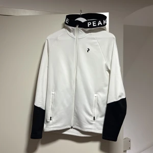 Vit jacka från Peak Performance - Snygg vit jacka från Peak Performance med svarta detaljer. Jackan har dragkedja framtill och två praktiska fickor med dragkedjor. Den har en hög krage med text och svarta ärmslut.