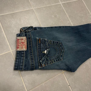 Mörkblå jeans från True Religion - Snygga mörkblå jeans från True Religion med klassisk femficksdesign och orange sömmar. Jeansen har en knappgylf och är i en bootcut-stil som ger en avslappnad look. Perfekta för en casual outfit.