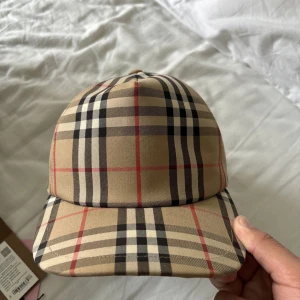Rutig keps från Burberry - Snygg rutig keps från Burberry i klassiskt beige, svart och rött mönster. Kepsen har justerbar passform och ett broderat märke på baksidan med texten 'Burberry London England'. Perfekt för en stilren look.