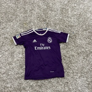 Real Madrid fotbollströja - Säljer en lila Real Madrid fotbollströja från Adidas med korta ärmar och vit text. Tröjan har 'Fly Emirates' tryckt på framsidan och 'Ronaldo 7' på baksidan. Perfekt för fans av laget!