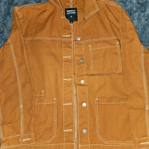 Brun jacka från Dr. Denim - Säljer en snygg brun jacka från Dr. Denim med vita sömmar och flera fickor. Jackan har knappar framtill och en stilren design som passar perfekt för höst och vår.