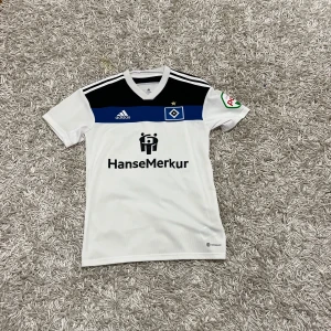 Hamburg fotbollströja - Säljer en vit fotbollströja från Adidas med svarta och blå detaljer. Tröjan har korta ärmar och klubbens logga på bröstet. Perfekt för fotbollsfans!