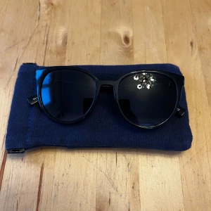 Svarta solglasögon från Le Specs - Snygga svarta solglasögon från Le Specs med en klassisk rundad form. Perfekta för soliga dagar och kommer med ett praktiskt fodral. Stilrena och tidlösa, dessa glasögon är ett måste för alla som älskar mode.