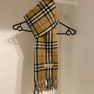 Burberry - Säljer denna Snygga och klassisk rutig halsduk från Burberry i beige. Tillverkad i mjuk lammull med fransar i ändarna. Perfekt för att hålla dig varm och stilren under kyliga dagar. Hör av er vid funderingar.