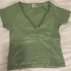 Grön v-ringad topp - Söt grön topp från Urban Outfitters med v-ringning och spetsdetaljer längs kanten. Toppen har en liten rosa ros framtill och korta ärmar. Perfekt för en avslappnad stil. Endast använd ett fåtal gånger!