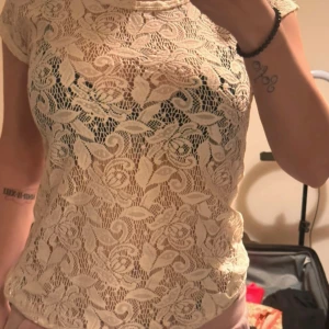 Vit spetsblus från Bella Ragazza - Säljer en elegant beige spetsblus från Bella Ragazza. Blusen har ett vackert blommönster och är kortärmad, perfekt för en stilren look.   