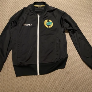 Svart träningsjacka från Craft - Säljer en svart träningsjacka från Craft med vit dragkedja och klubbmärke på bröstet. Texten 'BAJEN' i grönt på ryggen. Perfekt för sportiga aktiviteter.