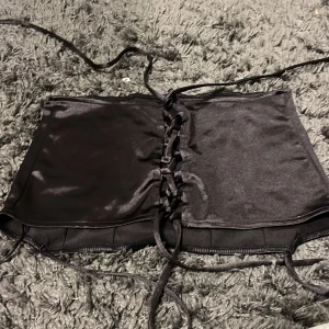 Svart korsett från H&M Divided - Snygg svart korsett från H&M Divided med snörning framtill. Perfekt för en stilren look. Tillverkad i ett glansigt material som ger en elegant touch.