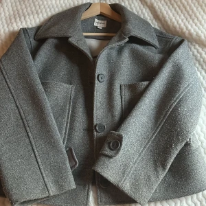 Grå kappa från Kappahl - Säljer en stilren grå kappa från Kappahl i storlek S. Jackan har en klassisk krage och knäppning med stora knappar framtill.