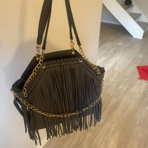 Mörkgrå axelväska med fransar från VILLA BAGS - Snygg mörkgrå axelväska med fransar och guldkedjedetaljer från VILLA BAGS. Köpt second hand men är helt i nyskick med lappen kvar.Väskan har en dragkedja och rymligt inre, perfekt för att bära med sig allt man behöver.  Har en liknade design som Stella McCartney väskor. 