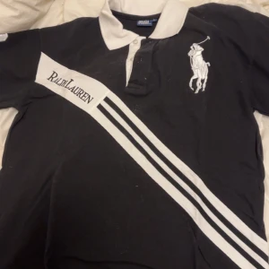Svart pikétröja från Ralph Lauren - Säljer en klassisk svart pikétröja från Ralph Lauren med vit krage och detaljer. Tröjan har en stor logga på bröstet och ett nummer på ärmen. Perfekt för en stilren look!