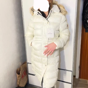 Vit dunjacka från Moncler - Säljer en snygg vit dunjacka från Moncler med huva och pälsdetaljer. Jackan har en lång design med dragkedja och knappar framtill. Perfekt för kalla dagar!