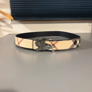 Burberry bälte - Snyggt burberry bälte med ett klassiskt rutigt mönster i beige, rött och svart. Bältet har en unik spänne i silver med en detaljerad design. Perfekt för att ge en stilren touch till din outfit.