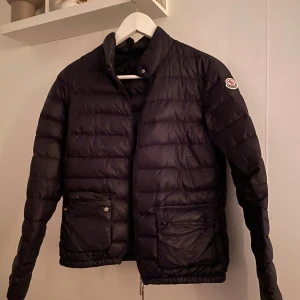 Svart dunjacka från Moncler - Snygg svart dunjacka från Moncler med klassisk design. Jackan har en hög krage och praktiska fickor med knappar. Perfekt för kyligare dagar och ger en stilren look. Lite suddiga bilder men fråga om fler och vad ni vill se ❤️🫶🏽 jackan är äkta och det som tillkommer kan ni se på bild 3 