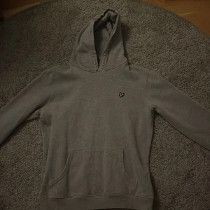 Grå hoodie från Lyle & Scott - Säljer en stilren grå hoodie från Lyle & Scott. Den har en klassisk design med en liten gul logga på bröstet och en praktisk magficka. Perfekt för en avslappnad look.
