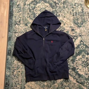 Blå hoodie från Polo Ralph Lauren - Säljer en stilren svart hoodie från Polo Ralph Lauren med dragkedja och den klassiska röda loggan på bröstet. Perfekt för en avslappnad stil. Den har en bekväm passform och är tillverkad i mjukt material.