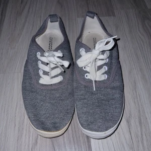 Grå sneakers från Divided - Säljer ett par grå sneakers från Divided i storlek 38. Skorna har vita snören och en vit sula. Perfekta för en avslappnad stil.
